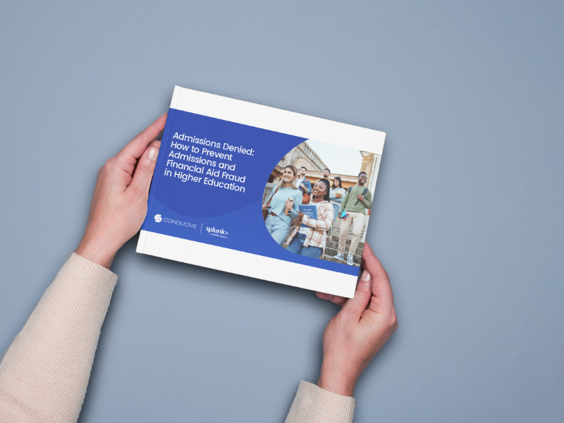 Hands holding a printed copy of the “Admissions Denied: How to Prevent Admissions and Financial Aid Fraud in Higher Education” e-book, featuring Conducive and Splunk logos, against a light blue background.
